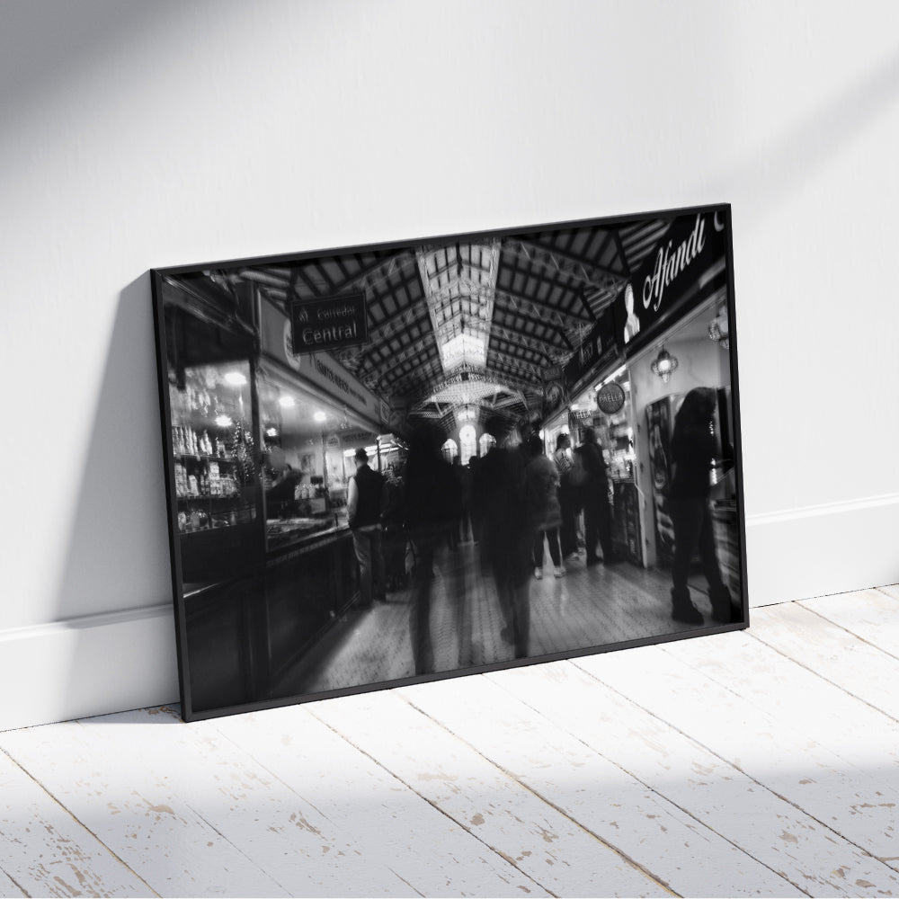 Fotografia in bianco e nero di un mercato coperto con persone in movimento tra le bancarelle illuminate. La struttura del soffitto presenta dettagli architettonici elaborati, con una finestra centrale luminosa che cattura l'attenzione. L'atmosfera è vivace ma sfocata, suggerendo dinamismo e attività.
