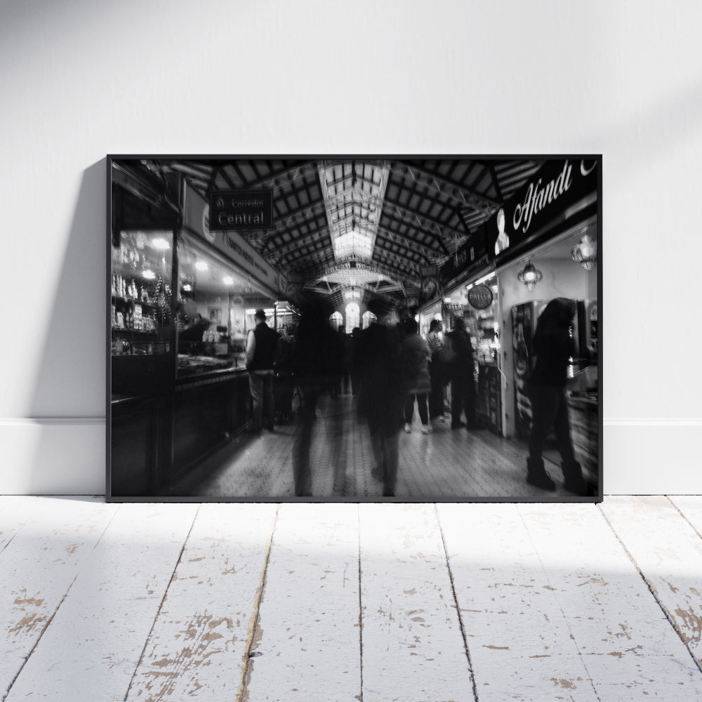 Fotografia in bianco e nero di un mercato coperto con persone in movimento tra le bancarelle illuminate. La struttura del soffitto presenta dettagli architettonici elaborati, con una finestra centrale luminosa che cattura l'attenzione. L'atmosfera è vivace ma sfocata, suggerendo dinamismo e attività.