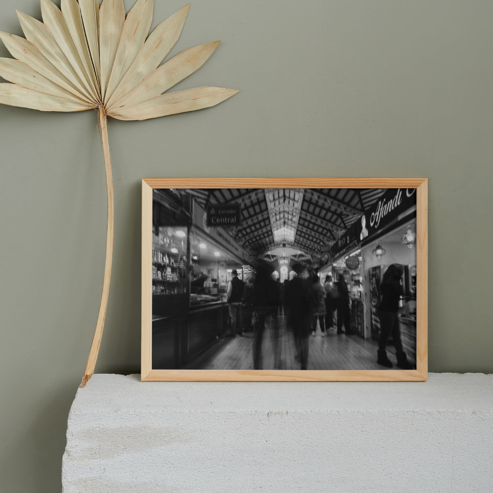 Fotografia in bianco e nero di un mercato coperto con persone in movimento tra le bancarelle illuminate. La struttura del soffitto presenta dettagli architettonici elaborati, con una finestra centrale luminosa che cattura l'attenzione. L'atmosfera è vivace ma sfocata, suggerendo dinamismo e attività.