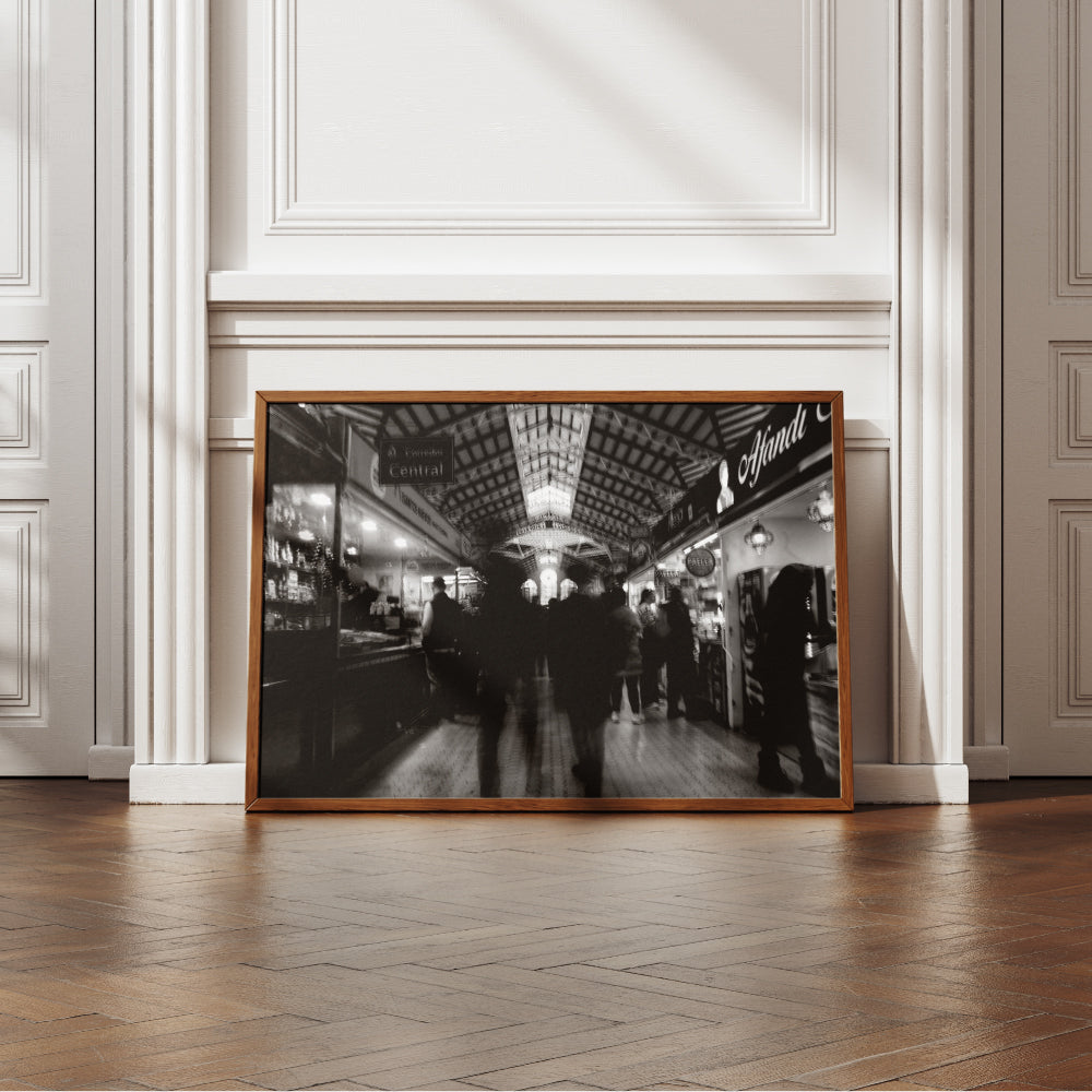 Fotografia in bianco e nero di un mercato coperto con persone in movimento tra le bancarelle illuminate. La struttura del soffitto presenta dettagli architettonici elaborati, con una finestra centrale luminosa che cattura l'attenzione. L'atmosfera è vivace ma sfocata, suggerendo dinamismo e attività.