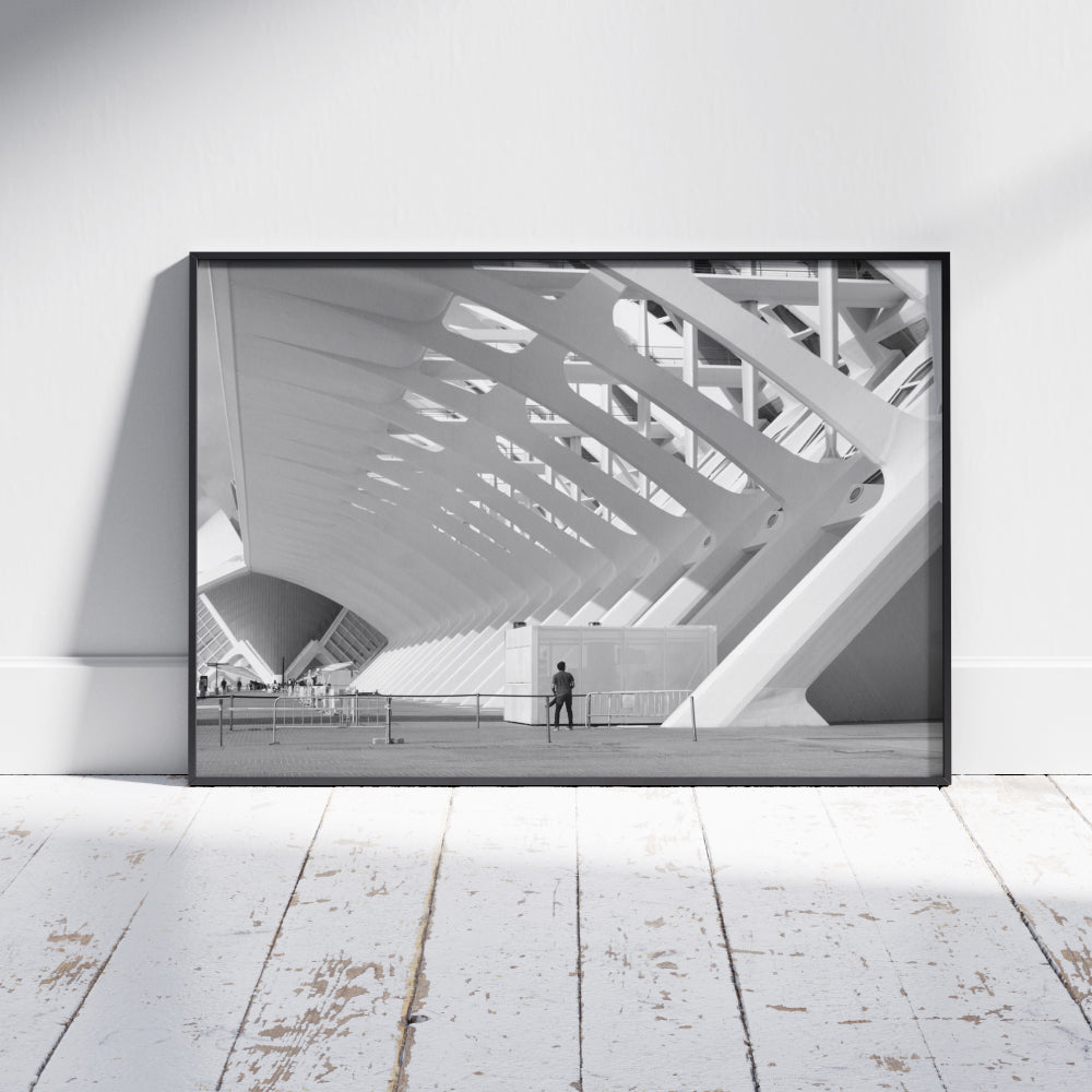 Struttura architettonica moderna con grandi arcate bianche e design geometrico, illuminata dalla luce del giorno. Una persona in piedi vicino a una recinzione guarda l'edificio, con piccoli gruppi di persone visibili in lontananza.