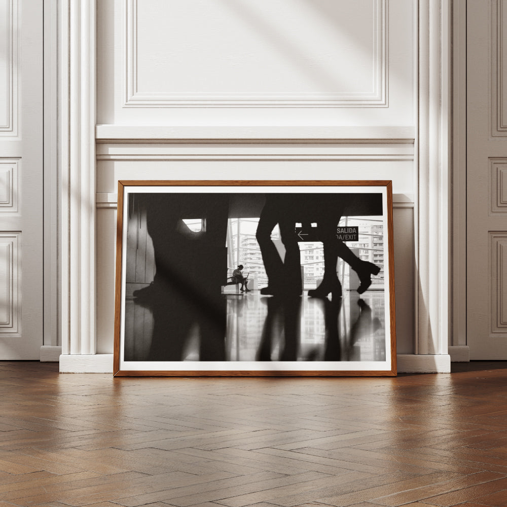 Scena in bianco e nero di una persona seduta su una panchina all'interno di un edificio moderno, visibile attraverso le silhouette sfocate di persone in movimento. Sullo sfondo, una vetrata mostra edifici urbani e un'uscita segnalata con un cartello 'Salida/Exit'.