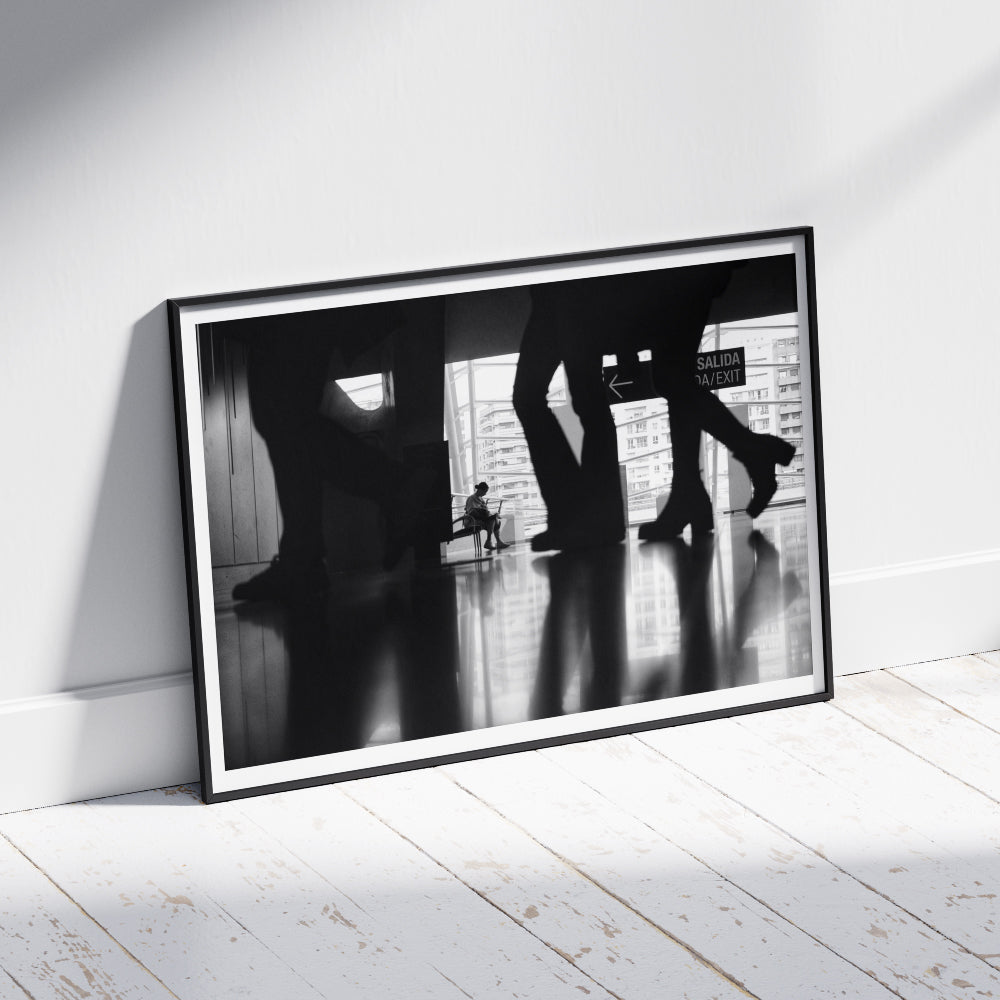 Scena in bianco e nero di una persona seduta su una panchina all'interno di un edificio moderno, visibile attraverso le silhouette sfocate di persone in movimento. Sullo sfondo, una vetrata mostra edifici urbani e un'uscita segnalata con un cartello 'Salida/Exit'.