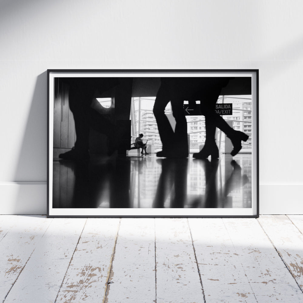 Scena in bianco e nero di una persona seduta su una panchina all'interno di un edificio moderno, visibile attraverso le silhouette sfocate di persone in movimento. Sullo sfondo, una vetrata mostra edifici urbani e un'uscita segnalata con un cartello 'Salida/Exit'.