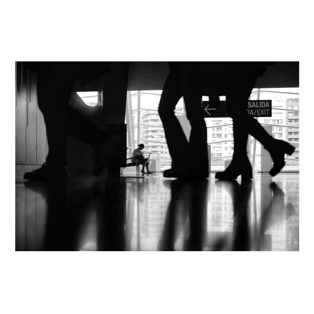 Scena in bianco e nero di una persona seduta su una panchina all'interno di un edificio moderno, visibile attraverso le silhouette sfocate di persone in movimento. Sullo sfondo, una vetrata mostra edifici urbani e un'uscita segnalata con un cartello 'Salida/Exit'.