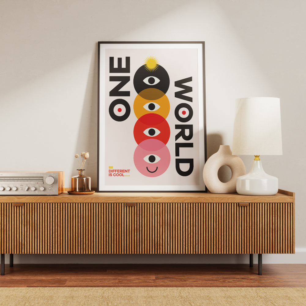 Poster con il testo 'One World' accompagnato da un design geometrico colorato con cerchi sovrapposti in nero, giallo, rosso e rosa, ciascuno contenente un occhio stilizzato e un volto sorridente nell'ultimo cerchio. In basso a sinistra la scritta 'Different is Cool'. Design moderno e simbolico su sfondo chiaro.