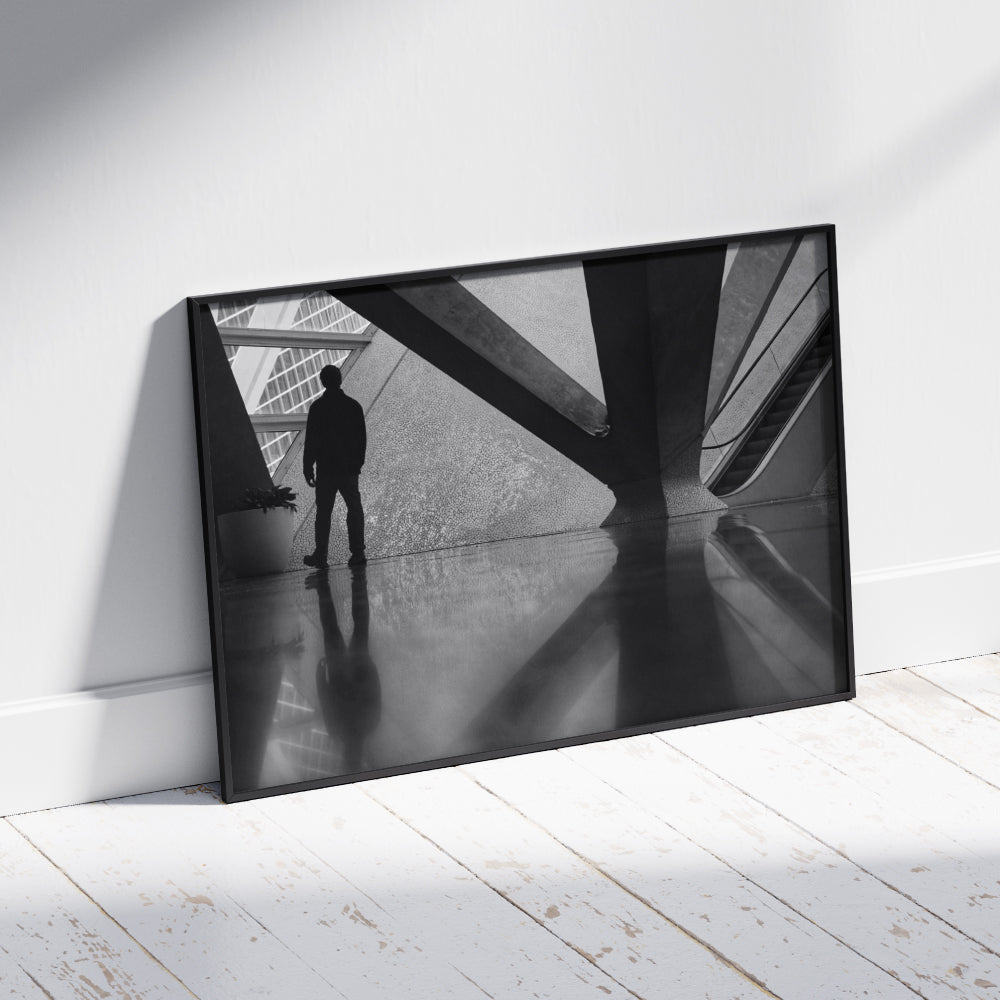 Silhouette di una persona in piedi davanti a una struttura architettonica moderna con dettagli geometrici e texture a mosaico. Sul lato destro si intravede una scala mobile, mentre il pavimento lucido riflette la figura e gli elementi circostanti.