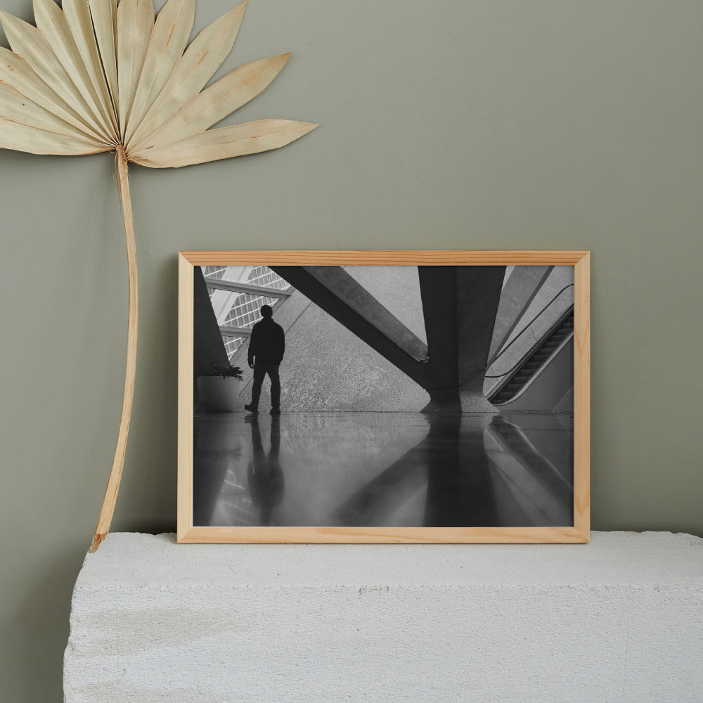 Silhouette di una persona in piedi davanti a una struttura architettonica moderna con dettagli geometrici e texture a mosaico. Sul lato destro si intravede una scala mobile, mentre il pavimento lucido riflette la figura e gli elementi circostanti.