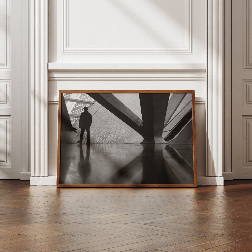 Silhouette di una persona in piedi davanti a una struttura architettonica moderna con dettagli geometrici e texture a mosaico. Sul lato destro si intravede una scala mobile, mentre il pavimento lucido riflette la figura e gli elementi circostanti.